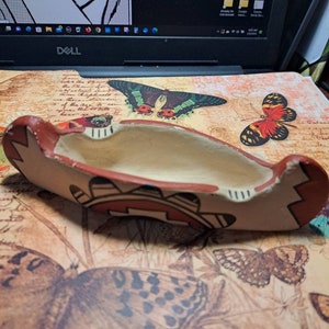 May include: A hand-painted, oblong ceramic ashtray with a cream-colored interior and a reddish-brown exterior. The ashtray features geometric designs in red, black, and white. The design is reminiscent of Native American art.