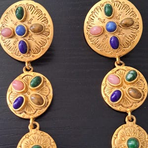 May include: Gold-toned dangle earrings with a circular design. Each earring features a series of colorful gemstones, including pink, green, blue, and brown. The earrings have an ornate, engraved pattern and a post-back closure.