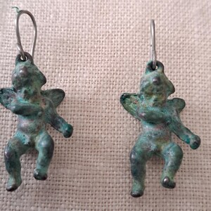 May include: A pair of antique-style earrings featuring small, weathered figures with outstretched arms and wings. The earrings have a green-blue patina and are suspended from silver-colored ear wires. The figures appear to be cherubs or similar mythological beings.