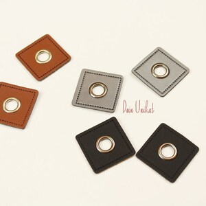 Può includere: Cinque toppe quadrate in pelle con occhielli metallici. Le toppe sono marroni, grigie e nere. Il testo "Dein Unikat" è visibile nell'immagine.