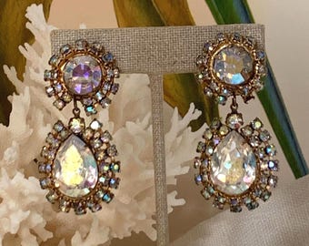 May include: A pair of drop earrings featuring a round top stone and a larger pear-shaped bottom stone, both with an iridescent finish. Each stone is surrounded by a halo of small, sparkling crystals set in a gold-toned metal frame.