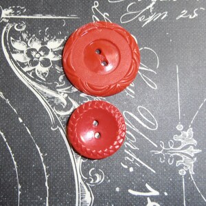 May include: Two red plastic buttons with a raised, braided design around the edge. The buttons have two holes each.