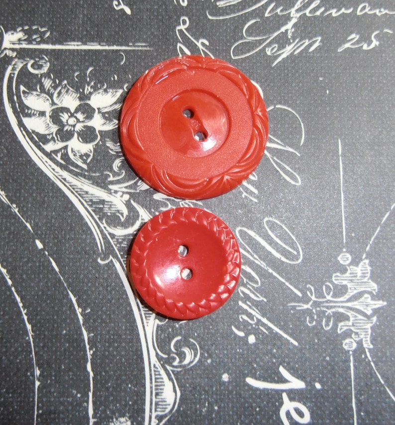 May include: Two red plastic buttons with a raised, braided design around the edge. The buttons have two holes each.