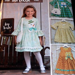 May include: A sewing pattern for a girl's dress with a snowman applique. The pattern includes three different dress styles: a green plaid dress with a bear applique, a yellow floral dress, and an orange polka dot dress with an owl applique. The pattern also includes a white fuzzy purse.