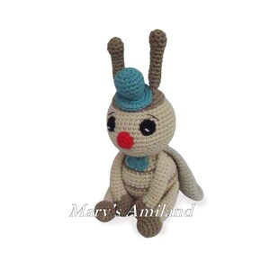 Può includere: Un insetto all'uncinetto beige e marrone con accenti blu e verdi. L'insetto indossa un cappello blu e ha un naso rosso. L'insetto è seduto con le gambe incrociate e le ali spiegate.