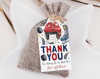 Puede incluir: Una bolsa de arpillera con una etiqueta de papel rectangular atada con cordel. La etiqueta muestra ilustraciones de equipo de hockey, el texto THANK YOU, For Hitting The Ice With Me! y el nombre Love, William en tinta roja y azul.