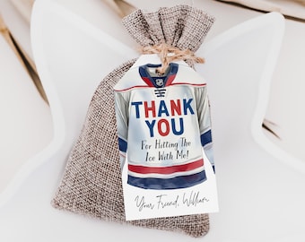 Puede incluir: Una bolsa de arpillera con cordón y una etiqueta de regalo de papel blanco atada con cuerda. La etiqueta presenta una ilustración de una camiseta de hockey con el texto THANK YOU For Hitting The Ice With Me! y la firma Your Friend, William.
