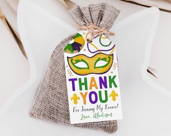 Peut inclure: Une pochette à cordon en toile de jute avec une étiquette en papier rectangulaire attachée par une ficelle. L'étiquette présente un masque de Mardi Gras, des perles et des fleurs de lys avec le texte THANK YOU For Joining My Krewe! Love, Madison.