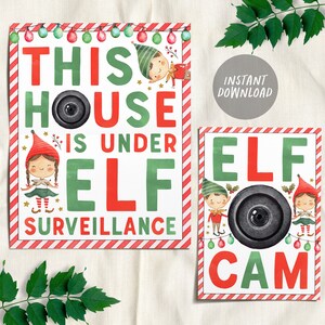 May include: Two printable signs with a red and white striped border. The first sign says "This house is under elf surveillance" with a cartoon elf and a camera. The second sign says "Elf Cam" with two cartoon elves and a camera.