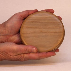 Puede incluir: Un objeto redondo de madera con un acabado marrón claro. El objeto se sostiene con dos manos, mostrando su superficie lisa y pulida. La veta de la madera es visible, agregando textura. Parece ser una tapa o un pequeño recipiente.
