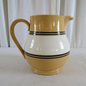 Può includere: Una brocca in ceramica con corpo arrotondato e manico curvo. La brocca è principalmente giallo senape, con una fascia bianca al centro e strisce nere. Ha un beccuccio per versare.