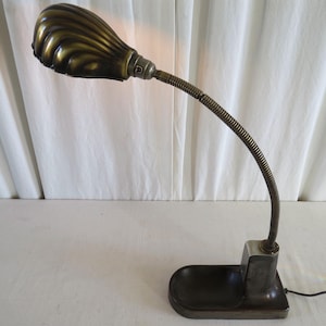 May include: An antique desk lamp with a brass-coloured, shell-shaped shade. The flexible, ribbed neck extends from a rectangular base with a small tray. The lamp is plugged into a wall outlet.