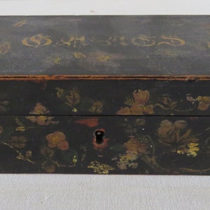 Könnte beinhalten: Eine rechteckige schwarze Box mit Scharnierdeckel. Die Box ist mit goldfarbenen Blumenmustern und dem Wort "SHORES" in goldfarbenen Buchstaben verziert. Ein Schlüsselloch ist auf der Vorderseite der Box sichtbar.