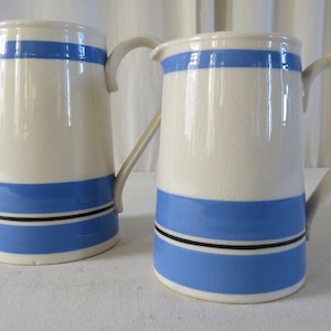 Puede incluir: Dos jarras de cerámica blanca con rayas azules y negras. Cada jarra tiene un asa curva y un pico vertedor. Las jarras son cilíndricas, con una base ancha y una parte superior más estrecha.