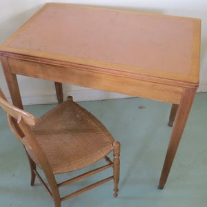 May include: A vintage wooden desk with a light brown top and four slender legs. A matching chair with a woven seat and curved back is positioned beside the desk. The desk and chair are made of light-colored wood.