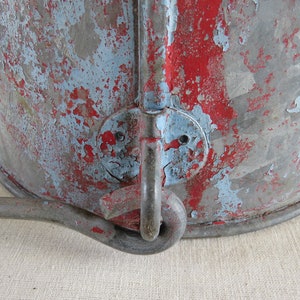 May include: Close-up of a vintage metal bucket with peeling paint. The bucket has a distressed appearance with red and blue paint over a grey metal base. A metal handle is attached to the side.