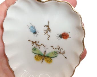 May include: A white porcelain trinket dish shaped like an apple with a scalloped edge and gold trim. The center features hand-painted illustrations of a blue beetle, a red ladybug, a yellow and green butterfly, and delicate brown vine motifs.