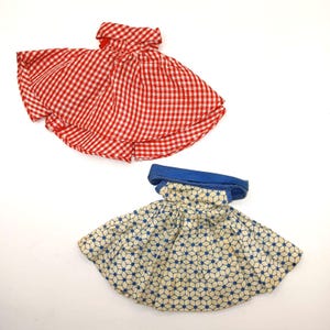 May include: Two doll skirts. One skirt features a red and white gingham checkered pattern with a folded waistband. The second skirt has a blue solid waistband and a white fabric base with a repeating blue geometric hexagon and dot pattern.