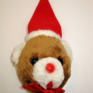 May include: A plush teddy bear head ornament featuring brown and white fur, wearing a red Santa hat with a green pom-pom on top. The bear has black plastic eyes, a round red plastic nose, and a red ribbon bow tied at the base of the chin.