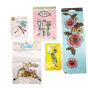 May include: A collection of five craft applique patches including a snowman with blue and red sequins, a cat illustration, a gold sequined deer, a green cartoon character on a yellow card, and a long floral embroidered patch with purple flowers.