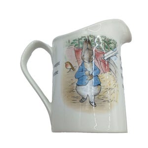 May include: A white ceramic creamer pitcher featuring a colorful illustration of Peter Rabbit wearing a blue jacket, standing near a small bird and potted plants. The surface includes printed text fragments that read went some and the.