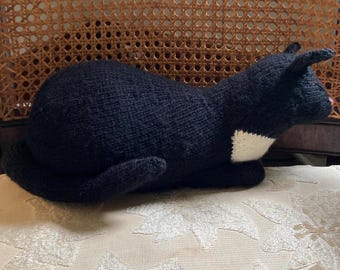 Puede incluir: Un juguete de peluche de gato negro hecho a mano con un parche blanco en el pecho, orejas y cola. El gato está acostado sobre una superficie estampada. La nariz del gato es rosa. El peluche está hecho de material de punto.