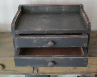 Op de afbeelding: Een kleine, donkergrijze houten doos met twee laden en een bovenste plank. De doos heeft een verweerde afwerking en is perfect voor het opbergen van kleine voorwerpen.