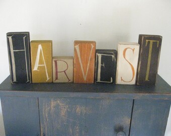 Op de afbeelding: Houten blokken letters die het woord "HARVEST" spellen in verschillende kleuren, waaronder zwart, geel, rood, oranje en wit. De letters zijn gerangschikt op een donkerblauw oppervlak.