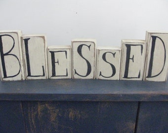 Op de afbeelding: Witte houten blokken die het woord "BLESSED" in zwarte letters spellen. De blokken zijn in een rij op een donkerblauw oppervlak gerangschikt.