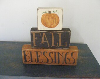 Op de afbeelding: Een driedelige houten blokkenset met de woorden "FALL BLESSINGS" in zwart op een bruine achtergrond. De bovenste blok heeft een geschilderde oranje pompoen met een groene steel.