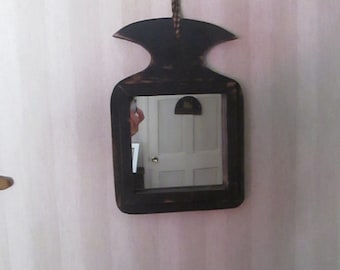 Op de afbeelding: Een zwarte houten spiegel met een eenvoudig ontwerp, hangend aan een witte muur. De spiegel heeft een rechthoekige vorm met een kleine, afgeronde bovenkant en een touwtje om op te hangen.
