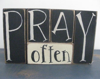 Op de afbeelding: Zwarte houten blokken die "PRAY often" spellen in witte verf. De blokken zijn in een rij gerangschikt met het woord "often" op het onderste blok.