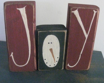 Op de afbeelding: Houten blokken met de tekst "JOY" en een sneeuwpop in het midden. De blokken "J" en "Y" zijn bordeauxrood met witte letters. Het sneeuwpopblok is zwart met een ovaal wit gezicht, een wortelneus en stipogen en -mond.