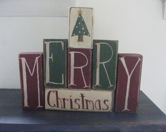Op de afbeelding: Een houten blokkenbord dat "Merry Christmas" spelt met een groene boom erop. De blokken zijn geschilderd in tinten rood en groen.
