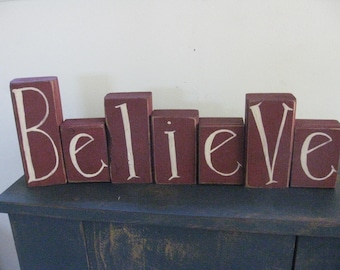 Op de afbeelding: Een houten blokkenbord dat het woord "Believe" spelt in witte verf op een bordeauxrode achtergrond.