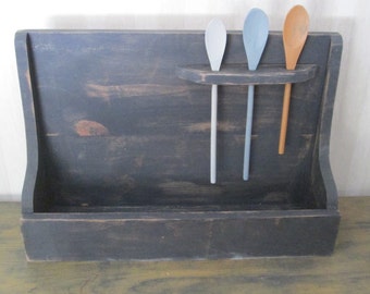 Op de afbeelding: Een zwarte houten bestekhouder met een plank om lepels aan te hangen. Drie houten lepels hangen aan de plank, één is lichtblauw, één is grijs en één is bruin.