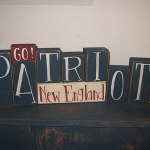 Könnte beinhalten: Holzbrettbuchstaben, die "GO! PATRIOTS New England" in weißer Farbe auf dunkelblauem Hintergrund buchstabieren. Der Block "GO!" ist rot.