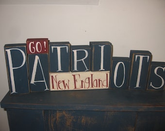 Op de afbeelding: Houten blokken letters die "GO! PATRIOTS New England" spellen in witte verf op een donkerblauwe achtergrond. De "GO!" blok is rood.