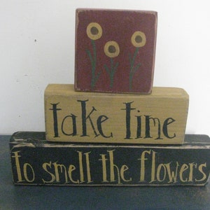 Może przedstawiać: Ułożone w stos drewniane klocki z napisami "take time" i "to smell the flowers" czarnymi literami. Górny klocek jest bordowy z żółtymi ilustracjami kwiatów. Rustykalny przedmiot dekoracyjny.