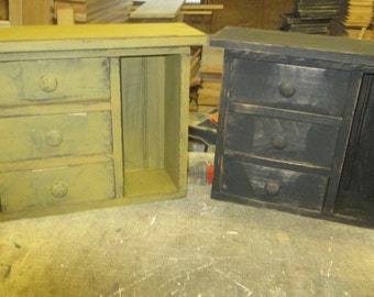 Op de afbeelding: Twee kleine houten kasten met elk drie laden. De kast links is lichtgeel geverfd en de kast rechts is zwart geverfd.