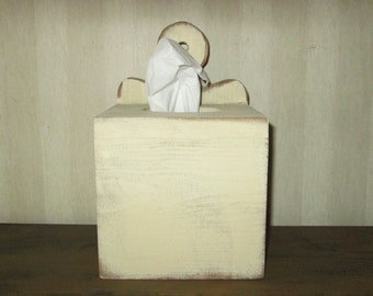 Op de afbeelding: Een crèmekleurige houten tissuebox met een verweerde afwerking. De doos heeft een vierkante vorm met een decoratieve bovenkant en een ronde opening voor het afgeven van tissues. Witte tissues zijn zichtbaar.