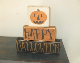Op de afbeelding: Een drielaags houten blokschild met de woorden "HAPPY HALLOWEEN" in zwart geschilderd op een bruine en zwarte achtergrond. De bovenste blok heeft een geschilderde oranje pompoen met een zwart gezicht.