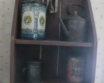 Op de afbeelding: Een houten plank met drie items erop. De bovenste plank heeft een blauw en wit blik met de woorden "Monarch Cocoa" erop, een kleine pop en een metalen waterkoker. De onderste plank heeft een metalen container en een rood en wit blik met de woorden "Rice Baking Powder" erop.