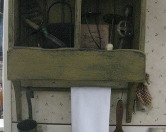Op de afbeelding: Een rustiek houten plank met twee vakken, gevuld met verschillende items, waaronder een kleine pop, een metalen zeef en een blikje met het opschrift "Cafe des Invalides". Een witte theedoek hangt aan de plank, met een rood hart en de tekst "Cold Nose Warm Heart".