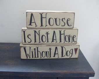 Op de afbeelding: Drie houten blokken gestapeld op elkaar met de woorden "A House Is Not A Home Without A Dog" in zwart geschilderd met een rood hart na het woord "Dog".