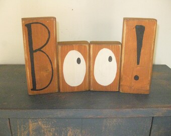 Op de afbeelding: Een houten blokkenbord dat "BOO!" spelt met witte oogjes en een zwart uitroepteken. De blokken zijn oranje geverfd.
