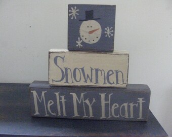 Op de afbeelding: Een driedelige houten blokkenset met een sneeuwmanontwerp op de bovenste blok, de middelste blok zegt "Snowmen" en de onderste blok zegt "Melt My Heart".