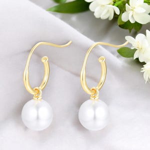以下が含まれることがあります： 金色のフープピアス。それぞれのフープから白い真珠が1つずつぶら下がっている。