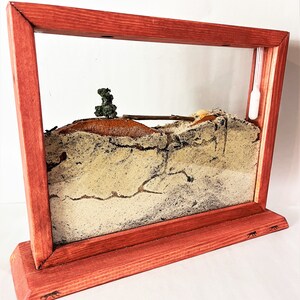 May include: A wooden ant farm with a red stained frame and a clear glass front. The ant farm is filled with sand and has a small tree and a bridge inside. There is a white plastic tube hanging from the top of the frame.