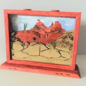 May include: A red wooden ant farm with a desert scene inside. The scene includes a cactus, red rock formations, and a sandy area with ant tunnels.
